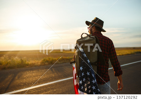 4th of July. American Flag. Traveler with the flag of America. The man in a hat, a backpack, a shirt and jeans. 137120292