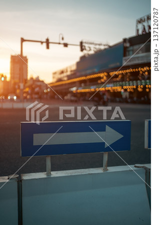 Directional arrow road sign at urban intersection during warm sunset at city atmosphere, Traffic guidance pointer on street with golden evening light and blurred city background 137120787