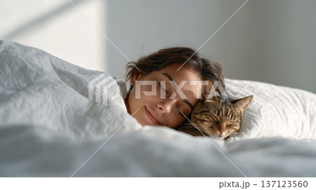 Woman and her tabby cat cuddling together, happily sleeping in a clean white bed with soft pillows and blankets, enjoying a peaceful morning nap or restful night 137123560