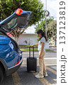 A teenager is standing next to a blue car with a suitcase in the trunk 137123819