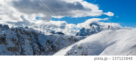 High resolution panoramic view of a ski mountaineer on a snowy ridge in the Dolomites with dramatic peaks and clouds, capturing winter adventure and vast alpine landscape. 137123848