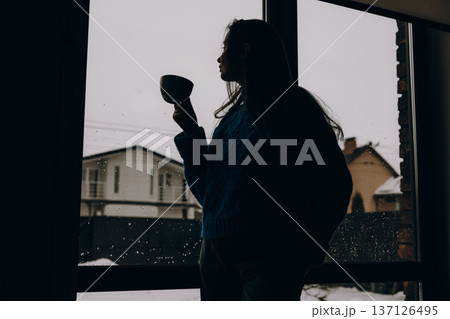 Silhouette of a young woman with a cup of coffee looking out of a window at a snowy backyard. Atmospheric moody indoor scene of solitude and winter comfort. Minimalist lifestyle photography background Silhouette of a young woman with a cup of coffee looking out of a window at a snowy backyard. Atmospheric moody indoor scene of solitude and winter comfort. Minimalist lifestyle photography background 137126495