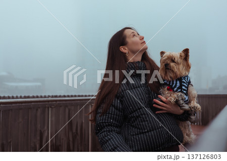 A beautiful young woman in a stylish puffer jacket holding a cute Yorkshire Terrier dog outdoors on a misty winter day. A bond between owner and pet. A beautiful young woman in a stylish puffer jacket holding a cute Yorkshire Terrier dog outdoors on a misty winter day. A bond between owner and pet. 137126803