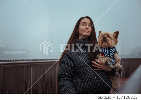 A beautiful young woman in a stylish puffer jacket holding a cute Yorkshire Terrier dog outdoors on a misty winter day. A bond between owner and pet. 137126815