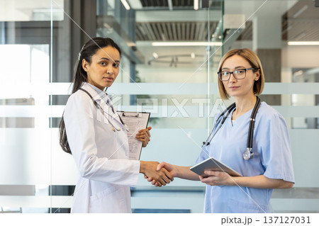 Two diverse female doctors, one holding a clipboard and another a tablet, are shaking hands and signifying agreement, collaboration, and partnership in a modern medical environment Two diverse female doctors, one holding a clipboard and another a tablet, are shaking hands and signifying agreement, collaboration, and partnership in a modern medical environment 137127031