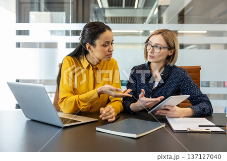 Two diverse businesswomen actively collaborating and communicating during a corporate meeting, one holding a digital tablet and the other gesturing, emphasizing teamwork and professional discussion Two diverse businesswomen actively collaborating and communicating during a corporate meeting, one holding a digital tablet and the other gesturing, emphasizing teamwork and professional discussion 137127040