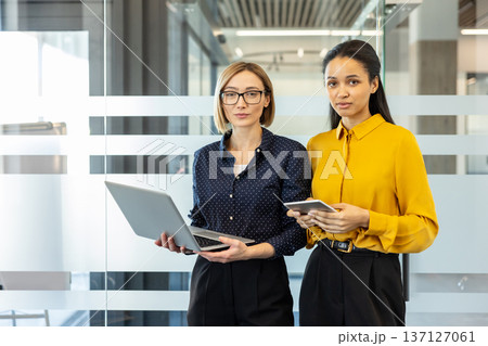 Two confident businesswomen, one holding a laptop and the other a tablet, standing together in a modern glass office, representing teamwork and professional partnership 137127061