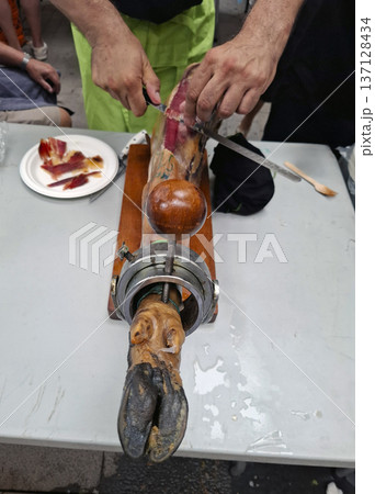 A professional carver skillfully cuts slices of Jamon Iberico on a wooden stand. The scene shows people nearby enjoying the flavors of Spain during a lively market day. 137128434