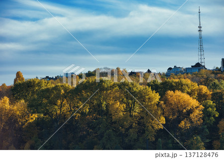 Autumn cityscape with colorful forest canopy in golden fall foliage, residential houses on a hilltop, and a tall radio tower under a blue sky with soft clouds. Autumn cityscape with colorful forest canopy in golden fall foliage, residential houses on a hilltop, and a tall radio tower under a blue sky with soft clouds. 137128476