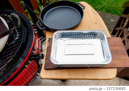 Ceramic grill with disposable aluminum trays and cast iron pan on side shelf outdoors Ceramic grill with disposable aluminum trays and cast iron pan on side shelf outdoors 137130685