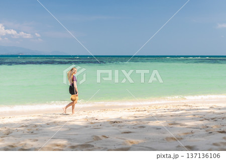 Young woman walking barefoot along tropical beach on Koh Samui, Thailand, holding straw hat and looking at turquoise sea under clear blue sky Young woman walking barefoot along tropical beach on Koh Samui, Thailand, holding straw hat and looking at turquoise sea under clear blue sky 137136106