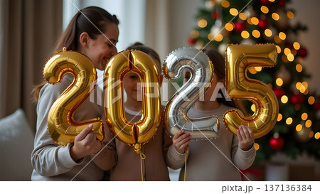 A family holds shiny golden balloons displaying 2025, standing in front of a decorated Christmas tree, radiating joy and excitement for the holidays and New Year celebrations 137136384