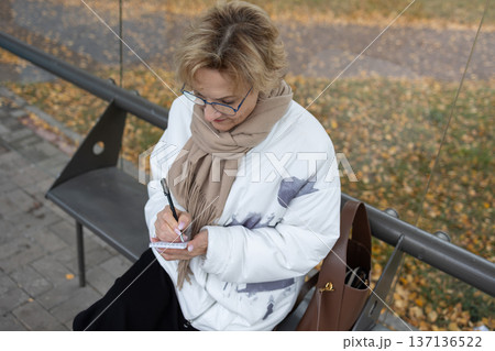 Mature woman with glasses holding notepad and pen outdoors near bench with autumn leaves on pavement. 137136522