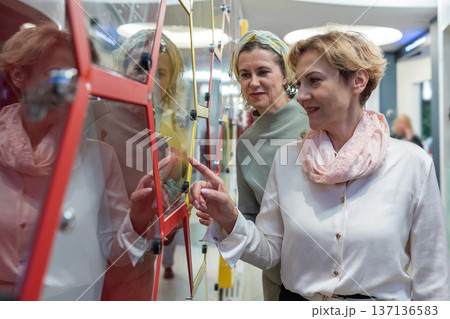 Women standing in gallery interior examining framed artwork and pointing during exhibition visit in quiet discussion. Women standing in gallery interior examining framed artwork and pointing during exhibition visit in quiet discussion. 137136583