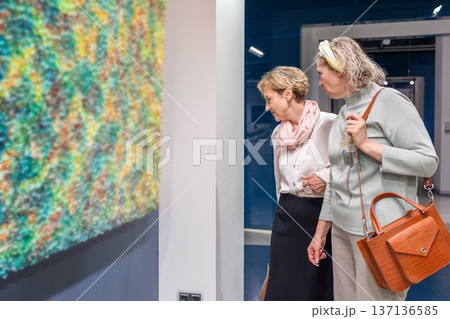 Women standing in gallery interior examining framed artwork and pointing during exhibition visit in quiet discussion. 137136585