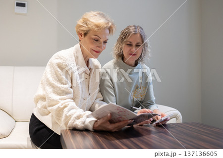 Two mature women sitting on light sofa examining open brochure on table discussing printed images in quiet interior space. 137136605