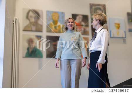 Two women appreciating contemporary art inside modern museum space sharing impressions and gestures while viewing framed paintings. Two women appreciating contemporary art inside modern museum space sharing impressions and gestures while viewing framed paintings. 137136617