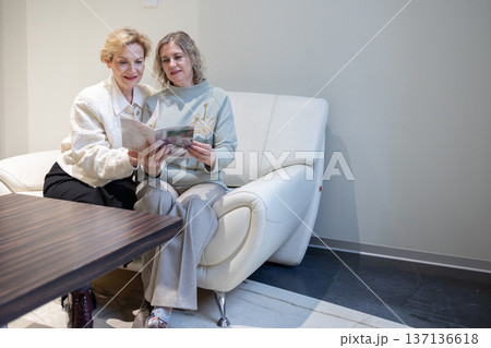 Collaboration. Women focused on catalog pages sharing opinions and pointing at photos while planning or selecting service in modern room. Collaboration. Women focused on catalog pages sharing opinions and pointing at photos while planning or selecting service in modern room. 137136618