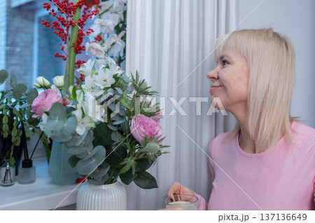 Sensory enjoyment. Woman in pink admiring and smelling flowers in vase, multiple bouquets on table, cozy interior, soft daylight. 137136649