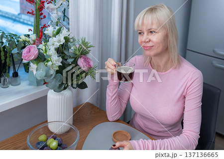 Mindful pause. Woman drinking hot coffee at home while sitting at table near fresh flowers soft daylight calm lifestyle mood. 137136665