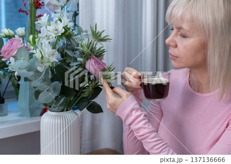 Domestic comfort moment. Woman drinking coffee indoors with floral decor natural light relaxed posture cozy everyday atmosphere. Domestic comfort moment. Woman drinking coffee indoors with floral decor natural light relaxed posture cozy everyday atmosphere. 137136666