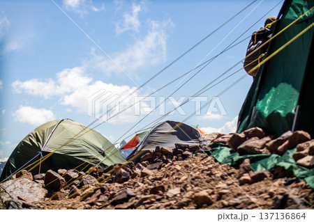 Mountain base camp with several colorful expedition tents pitched on rocky terrain under a blue sky with clouds. 137136864