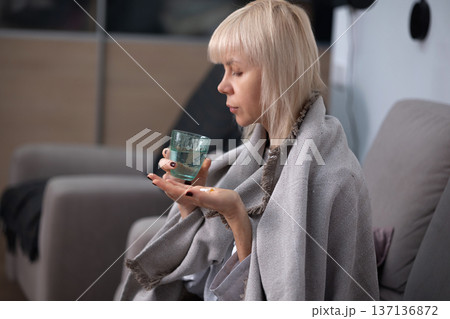 Medication intake. Blonde woman wrapped in blanket sitting on sofa holding pills in palm preparing to take medicine indoors during illness recovery. 137136872