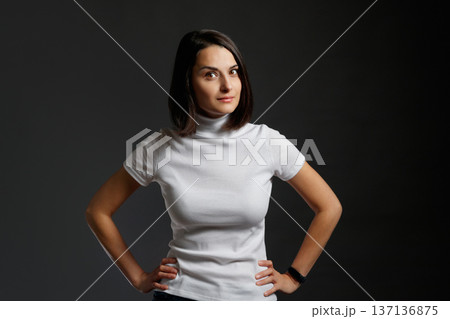 Portrait of a confident young woman in white turtleneck standing with her hands on hips and looking at camera. 137136875