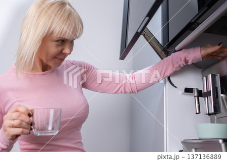 Coffee preparation. Adult blonde woman operating espresso machine holding empty glass cup in bright kitchen interior with focus on routine Coffee preparation. Adult blonde woman operating espresso machine holding empty glass cup in bright kitchen interior with focus on routine 137136889