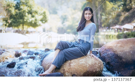 Young woman sitting on a boulder by a shallow river 137137367