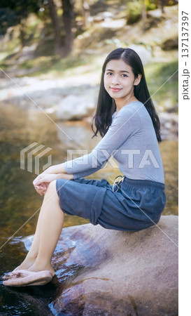 Smiling young woman sitting on riverside rock with feet near water, outdoor nature portrait with soft sunlight and shallow depth of field. Smiling young woman sitting on riverside rock with feet near water, outdoor nature portrait with soft sunlight and shallow depth of field. 137137397