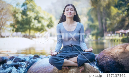 Young woman meditating cross-legged on rock by a calm river in sunny park, eyes closed, relaxed Young woman meditating cross-legged on rock by a calm river in sunny park, eyes closed, relaxed 137137561