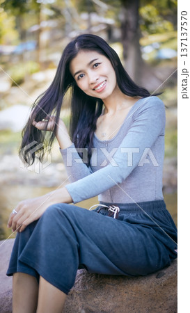 Smiling young woman with long dark hair sitting outdoors on a rock in a park Smiling young woman with long dark hair sitting outdoors on a rock in a park 137137570