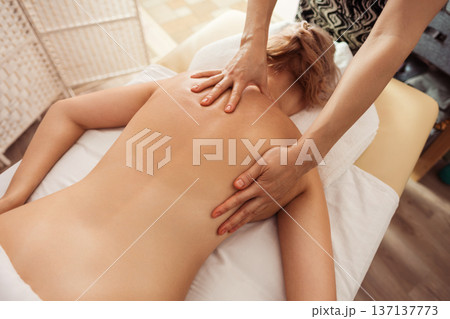 Overhead view of a spa therapist giving a soothing back massage to a young blonde woman on a white towel, warm soft light and neutral tones creating a calm wellness mood. 137137773
