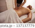 Spa therapist massaging the lower back of a young woman lying under a white sheet, warm natural light on smooth skin, wicker partition and soft textiles creating tranquil wellness atmosphere. 137137779