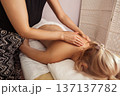 Relaxing spa massage with young blonde woman lying face down on white towel, receiving soothing back treatment from a therapist in patterned pants under warm, soft indoor light 137137782