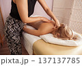 Relaxing spa massage with young blonde woman lying face down on white towel, receiving soothing back treatment from a therapist in patterned pants under warm, soft indoor light 137137785