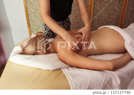 Close-up view of professional massage therapist performing back massage on a blonde woman lying on table, in spa room with warm light, calm tones, and relaxing atmosphere. Close-up view of professional massage therapist performing back massage on a blonde woman lying on table, in spa room with warm light, calm tones, and relaxing atmosphere. 137137790