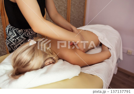 Close-up view of professional massage therapist performing back massage on a blonde woman lying on table, in spa room with warm light, calm tones, and relaxing atmosphere. 137137791