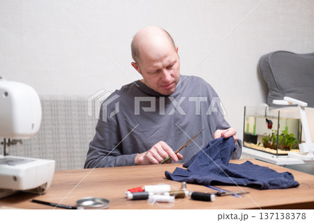 A bald man in his forties wearing a grey T-shirt sits at a table and cuts fabric with sewing scissors. 137138378