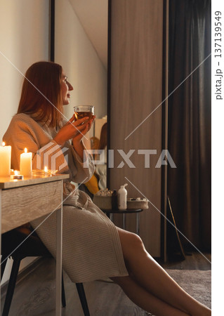 Woman in bathrobe enjoying hot tea illuminated by flickering candles, finding peace and tranquility. Wellness and spa evening 137139549