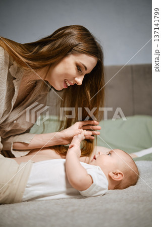 Mother interacts with baby on bed in home setting during daylight hours 137141799