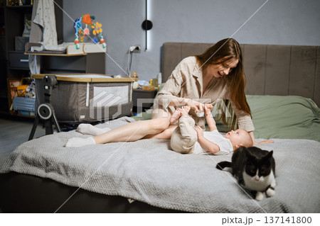 Mother plays with baby on the bed while cat walks around in a bedroom during the day 137141800