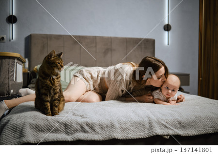 Mother and baby spend time together on bed with cat nearby in cozy bedroom setting Mother and baby spend time together on bed with cat nearby in cozy bedroom setting 137141801