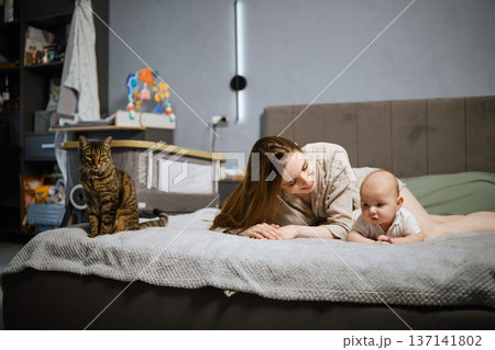 Mother and baby play together on bed with cat nearby in cozy room during daytime 137141802