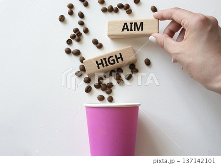 Aim High symbol. Concept word Aim High on wooden blocks. Beautiful white background with coffee cup. Businessman hand. Business and Aim High concept. Copy space 137142101