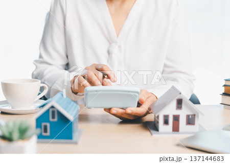 Mortgage and real estate budgeting concept with consultant using calculator beside miniature homes, coffee cup and paperwork on bright desk, symbolizing home loan planning and insurance. 137144683