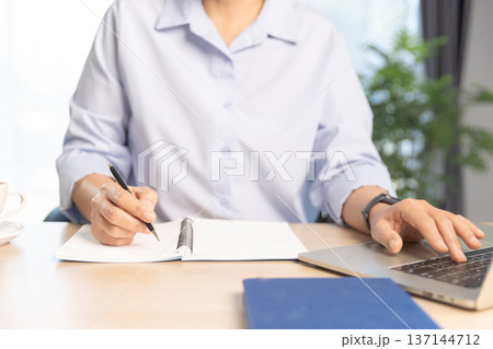 Business professional working on laptop at modern office desk with documents, smartphone and stationery, bright daylight by window. Concept of productivity, remote work and digital management. 137144712