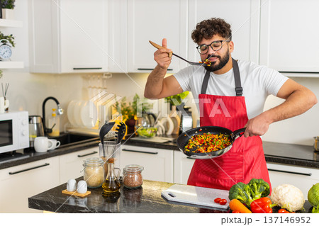 Arabian young adult man tasting vegetables from pan in kitchen cooking healthy dinner recipe at home Arabian young adult man tasting vegetables from pan in kitchen cooking healthy dinner recipe at home 137146952