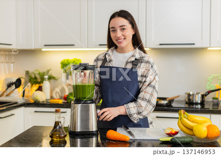 Young woman starts blender for green smoothie with spinach, banana, apple and carrot in kitchen 137146953